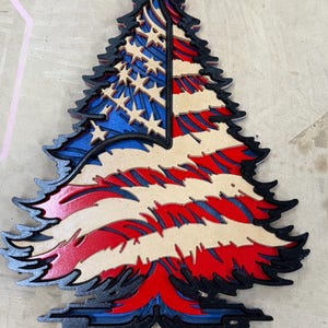 May include: A layered wooden tree decoration featuring the American flag design. The tree is painted with red, white, and blue colors, with stars on the blue section. The edges are outlined in black.