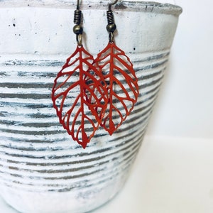 Puede incluir: Un par de pendientes de hoja naranja con un diseño delicado e intrincado. Los pendientes están hechos de metal y tienen un gancho de color bronce.