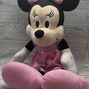 Pode incluir: Uma boneca de pel&uacute;cia da Minnie Mouse, com cabe&ccedil;a preta, orelhas grandes e la&ccedil;o rosa com bolinhas brancas. Ela usa um vestido rosa com bolinhas brancas e tem p&eacute;s rosa. A boneca &eacute; macia e fofinha.