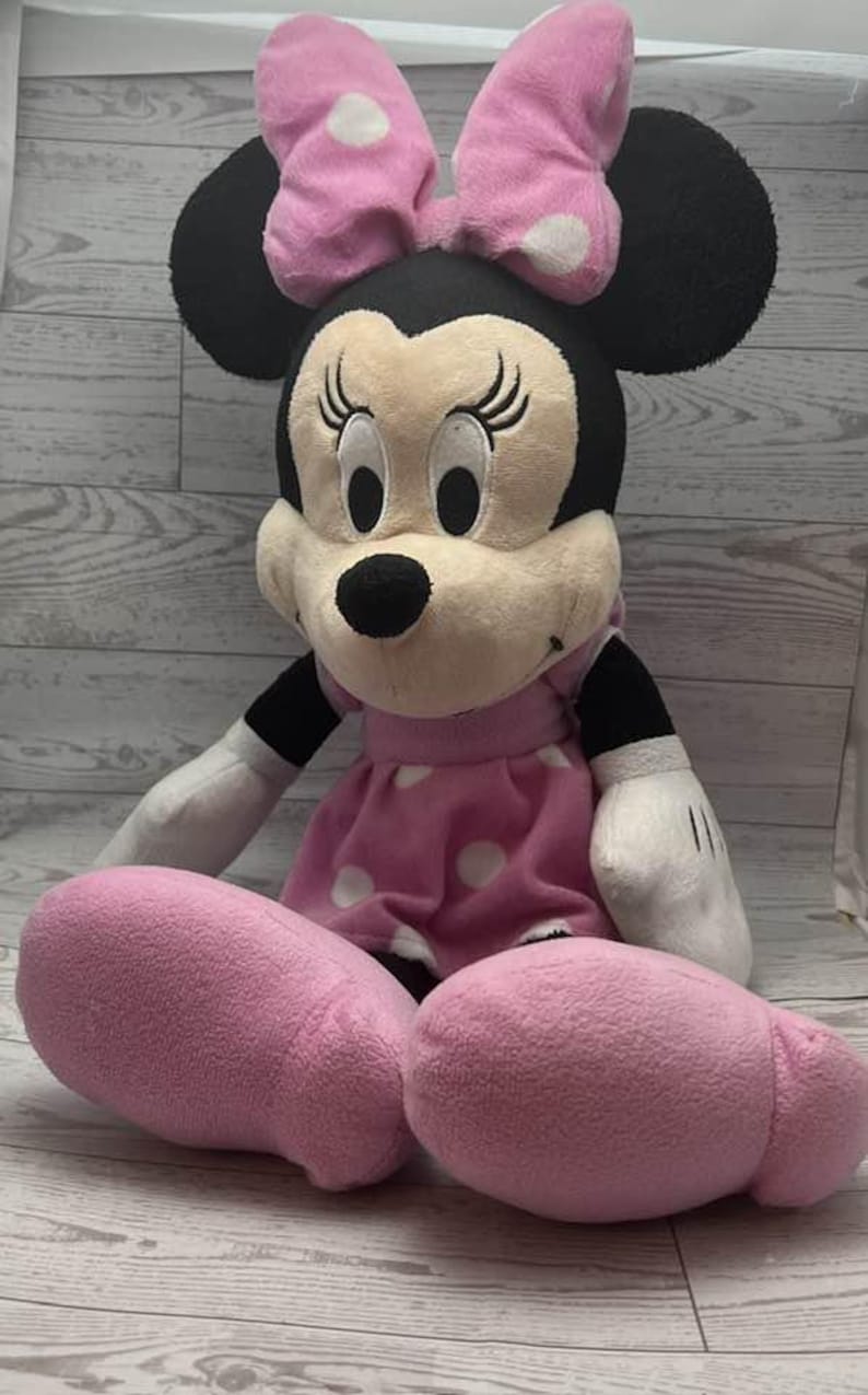 Pode incluir: Uma boneca de pel&uacute;cia da Minnie Mouse, com cabe&ccedil;a preta, orelhas grandes e la&ccedil;o rosa com bolinhas brancas. Ela usa um vestido rosa com bolinhas brancas e tem p&eacute;s rosa. A boneca &eacute; macia e fofinha.