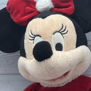 Puede incluir: Mu&ntilde;eca de peluche de Minnie Mouse con lazo y vestido rojos. La mu&ntilde;eca tiene orejas negras, nariz negra y cara color crema. El lazo tiene un pomp&oacute;n blanco en el centro. La mu&ntilde;eca lleva un vestido rojo con ribete blanco.