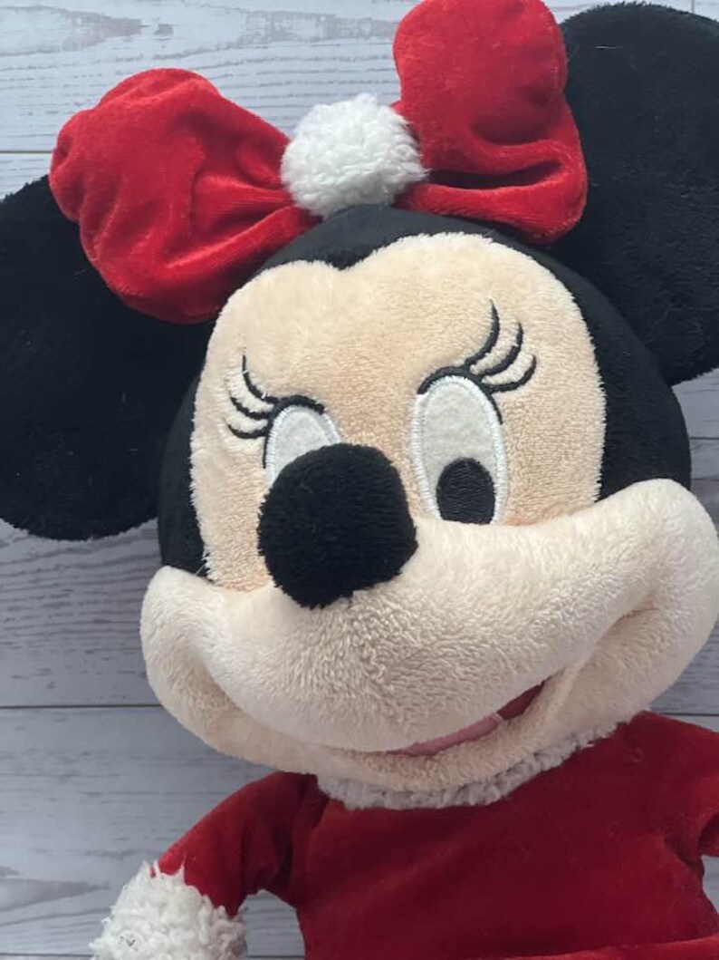 Puede incluir: Mu&ntilde;eca de peluche de Minnie Mouse con lazo y vestido rojos. La mu&ntilde;eca tiene orejas negras, nariz negra y cara color crema. El lazo tiene un pomp&oacute;n blanco en el centro. La mu&ntilde;eca lleva un vestido rojo con ribete blanco.