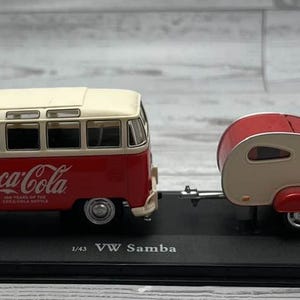 May include: A red and cream-colored VW Samba bus model with a matching teardrop trailer. The bus features the Coca-Cola logo and text. The model is displayed on a black base, with the text "1/43 VW Samba".