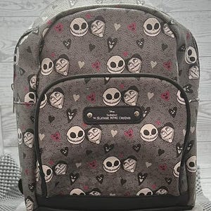 Puede incluir: Una mochila gris con un patr&oacute;n repetido de caras de Jack Skellington y otros personajes de "Pesadilla antes de Navidad". La mochila tiene ribetes negros y una peque&ntilde;a placa con el t&iacute;tulo de la pel&iacute;cula.