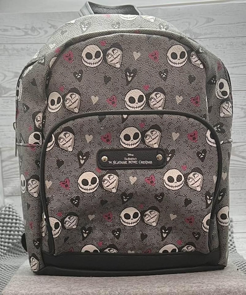 Puede incluir: Una mochila gris con un patr&oacute;n repetido de caras de Jack Skellington y otros personajes de "Pesadilla antes de Navidad". La mochila tiene ribetes negros y una peque&ntilde;a placa con el t&iacute;tulo de la pel&iacute;cula.