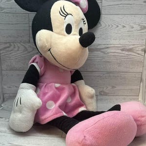 Pode incluir: Uma boneca de pel&uacute;cia da Minnie Mouse, com orelhas pretas, um nariz preto e um rosto sorridente. Ela usa um vestido rosa com bolinhas brancas e um la&ccedil;o combinando. Suas m&atilde;os e p&eacute;s s&atilde;o rosa.