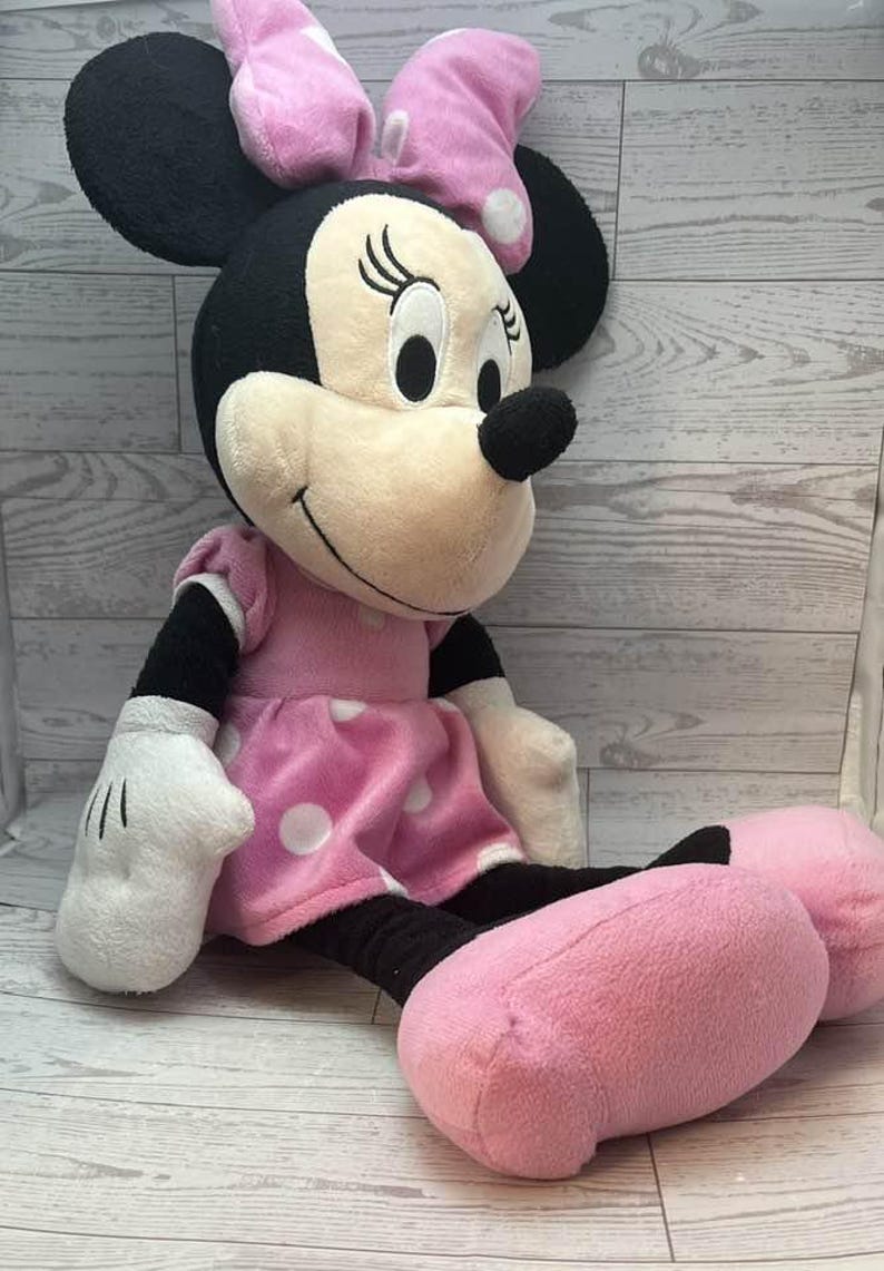 Pode incluir: Uma boneca de pel&uacute;cia da Minnie Mouse, com orelhas pretas, um nariz preto e um rosto sorridente. Ela usa um vestido rosa com bolinhas brancas e um la&ccedil;o combinando. Suas m&atilde;os e p&eacute;s s&atilde;o rosa.