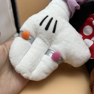 May include: A white plush glove with three black lines on the palm. The glove has a silver ring with an orange pom-pom and another silver ring with a pink pom-pom. Part of a red and white polka dot bow is visible.