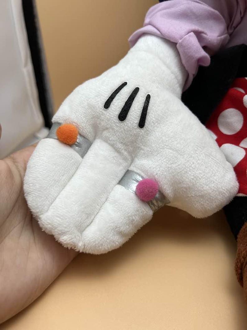 May include: A white plush glove with three black lines on the palm. The glove has a silver ring with an orange pom-pom and another silver ring with a pink pom-pom. Part of a red and white polka dot bow is visible.