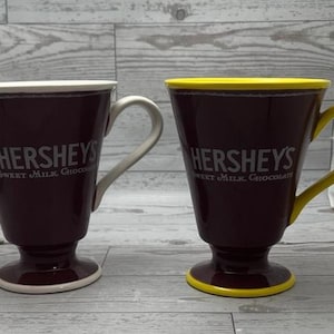 May include: Two Hershey's Sweet Milk Chocolate mugs. The mugs are dark brown with white text and handles. One mug has a white rim and handle, the other has a yellow rim and handle. The mugs have a pedestal base.