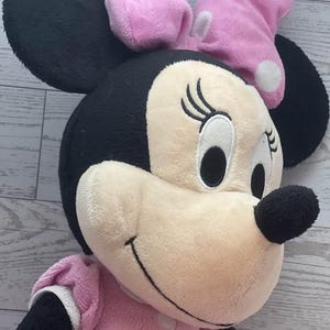 Pode incluir: Uma boneca de pel&uacute;cia da Minnie Mouse com orelhas pretas, cabe&ccedil;a preta e rosto bege claro. A boneca usa um vestido rosa e um la&ccedil;o rosa com bolinhas brancas. A boneca tem olhos pretos e nariz preto.