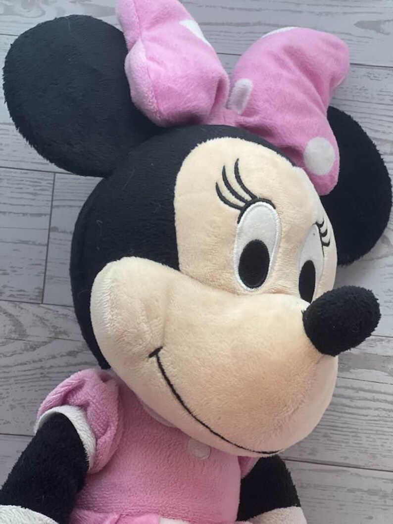 Pode incluir: Uma boneca de pel&uacute;cia da Minnie Mouse com orelhas pretas, cabe&ccedil;a preta e rosto bege claro. A boneca usa um vestido rosa e um la&ccedil;o rosa com bolinhas brancas. A boneca tem olhos pretos e nariz preto.