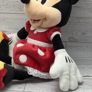 May include: A plush Minnie Mouse doll. The doll wears a red dress with white polka dots and a white lace trim. Minnie's bow is red, and her gloves are white with black lines. The doll has black ears and a tan face.