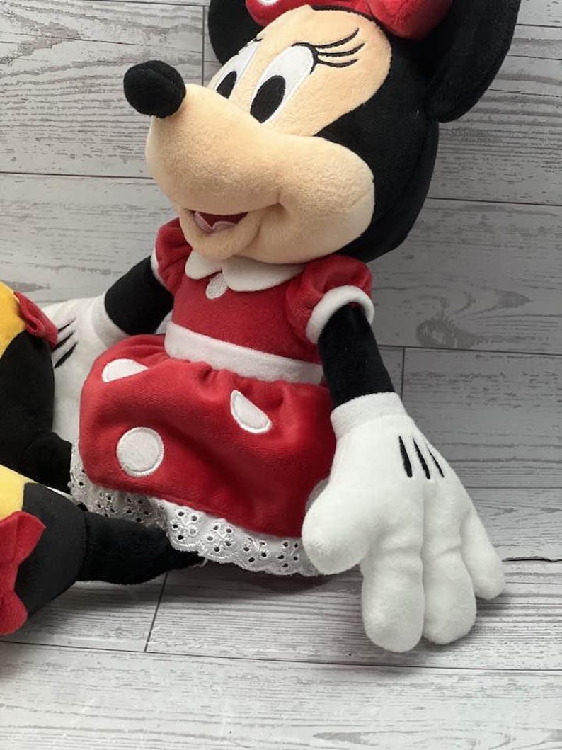 May include: A plush Minnie Mouse doll. The doll wears a red dress with white polka dots and a white lace trim. Minnie's bow is red, and her gloves are white with black lines. The doll has black ears and a tan face.
