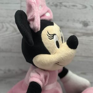 May include: A plush Minnie Mouse doll, featuring a black head and ears, a light pink bow, and a light pink dress. The doll has a beige face with embroidered eyes and a black nose. The doll's arms are white with black cuffs.