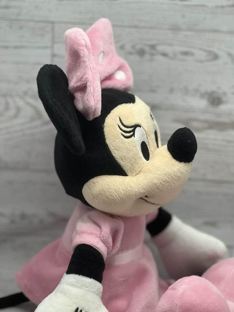 May include: A plush Minnie Mouse doll, featuring a black head and ears, a light pink bow, and a light pink dress. The doll has a beige face with embroidered eyes and a black nose. The doll's arms are white with black cuffs.