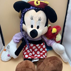 May include: A plush Minnie Mouse doll dressed as a pirate. She wears a red and gold pirate hat with a white feather, a purple bandana, and a red skirt with white polka dots. She has a red parrot on her arm and brown boots.