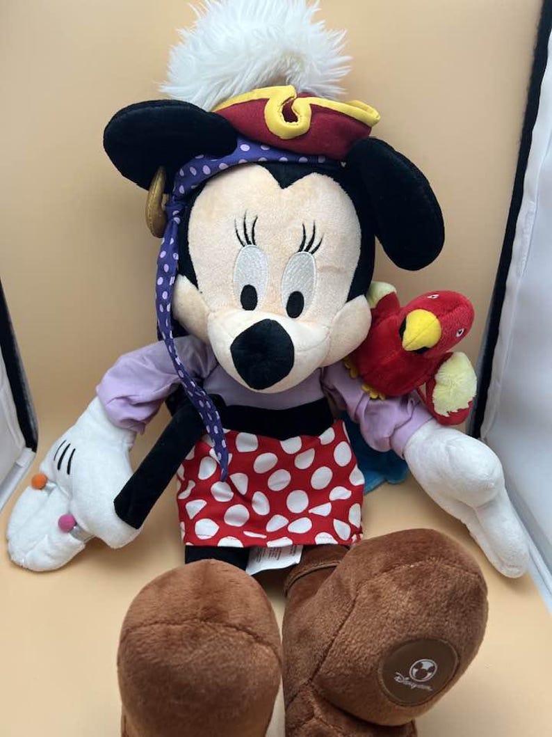 May include: A plush Minnie Mouse doll dressed as a pirate. She wears a red and gold pirate hat with a white feather, a purple bandana, and a red skirt with white polka dots. She has a red parrot on her arm and brown boots.