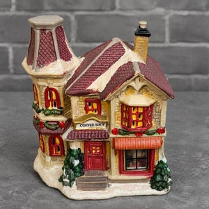 May include: A decorative ceramic coffee shop figurine with a red roof, chimney, and door. The building has multiple windows with red trim and a sign that reads "Coffee Shop". The base is covered in a dusting of white snow.
