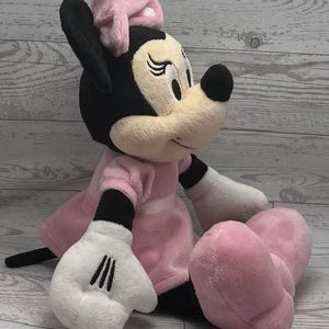 May include: A plush Minnie Mouse doll, featuring a black head and ears, a cream-colored face, and a pink bow. She wears a pink dress and shoes, with white gloves. The doll is sitting down.