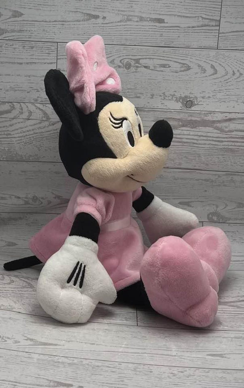 May include: A plush Minnie Mouse doll, featuring a black head and ears, a cream-colored face, and a pink bow. She wears a pink dress and shoes, with white gloves. The doll is sitting down.
