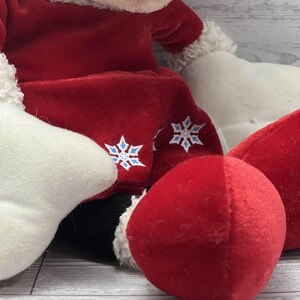 Puede incluir: Un peluche con un atuendo rojo con adornos blancos y detalles de copos de nieve. El juguete tiene una nariz roja y detalles en blanco y negro. El peluche es suave y parece ser un art&iacute;culo tem&aacute;tico de vacaciones.