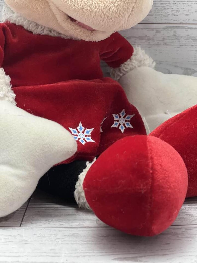 Puede incluir: Un peluche con un atuendo rojo con adornos blancos y detalles de copos de nieve. El juguete tiene una nariz roja y detalles en blanco y negro. El peluche es suave y parece ser un art&iacute;culo tem&aacute;tico de vacaciones.