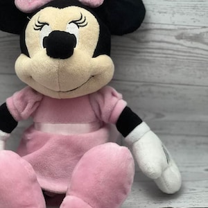 May include: A plush Minnie Mouse doll in a seated position. The doll features a pink dress, bow, and oversized feet. The bow has white polka dots. The doll has black ears, a black nose, and white gloves.