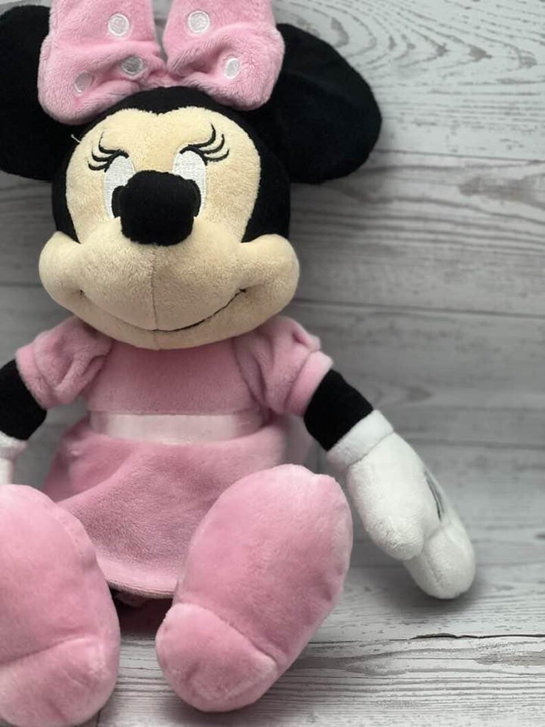 May include: A plush Minnie Mouse doll in a seated position. The doll features a pink dress, bow, and oversized feet. The bow has white polka dots. The doll has black ears, a black nose, and white gloves.