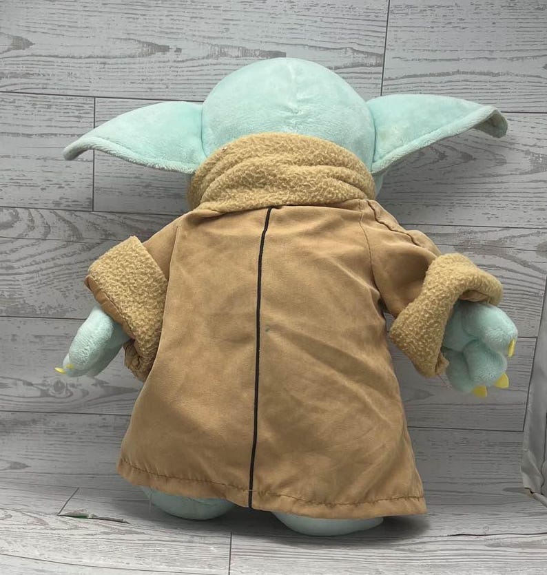 May include: A plush toy of a character with large ears, light green skin, and a tan coat with a fuzzy collar and cuffs. The toy is a soft, cuddly version of a popular character from a science fiction franchise.