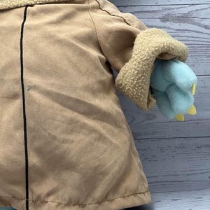 May include: A plush toy of a character from a popular science fiction series. The toy has a tan coat, a light blue face and hands, and a beige collar. The hands have yellow claws. The toy is on a light blue surface.