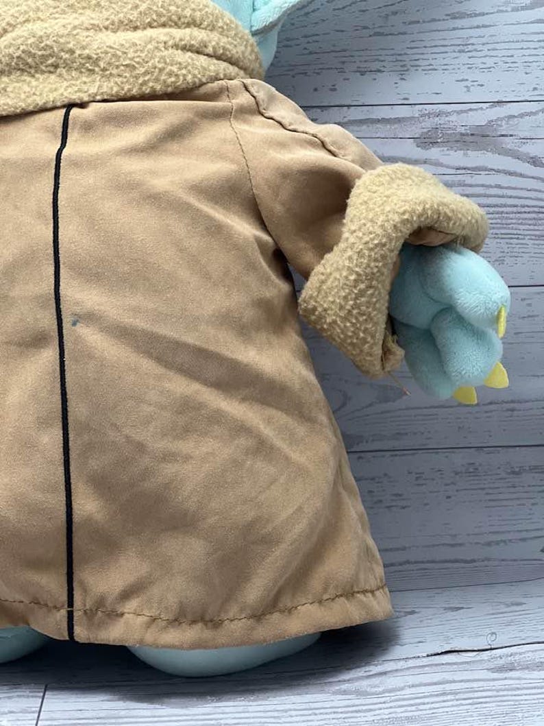 May include: A plush toy of a character from a popular science fiction series. The toy has a tan coat, a light blue face and hands, and a beige collar. The hands have yellow claws. The toy is on a light blue surface.