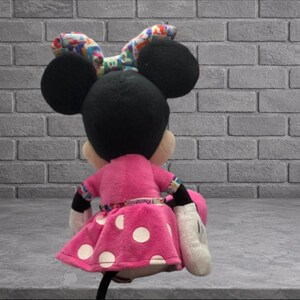 May include: A plush Minnie Mouse doll, viewed from the back, with a pink dress featuring white polka dots. The doll has black ears and a colorful bow. The doll is holding a pink purse and wearing white gloves.