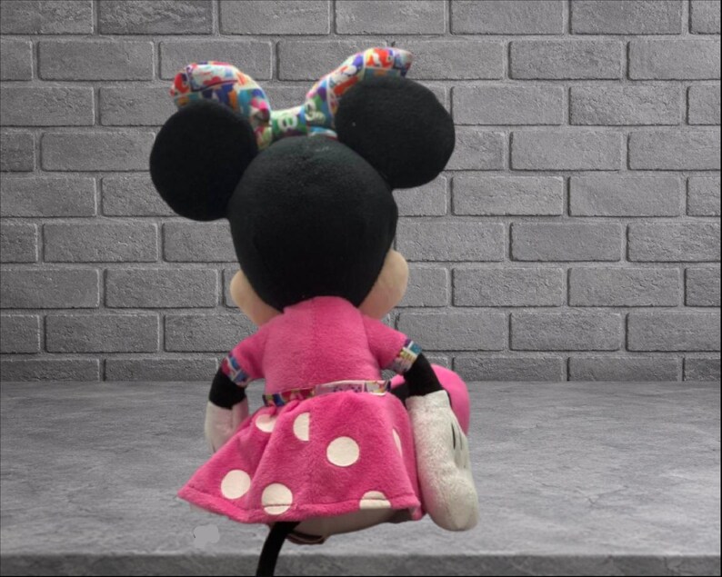 May include: A plush Minnie Mouse doll, viewed from the back, with a pink dress featuring white polka dots. The doll has black ears and a colorful bow. The doll is holding a pink purse and wearing white gloves.