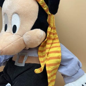 May include: A plush Mickey Mouse toy dressed as a pirate. The toy has a tan face, black ears, and is wearing a yellow and orange striped bandana. It is wearing a blue shirt, black pants, and a brown belt.