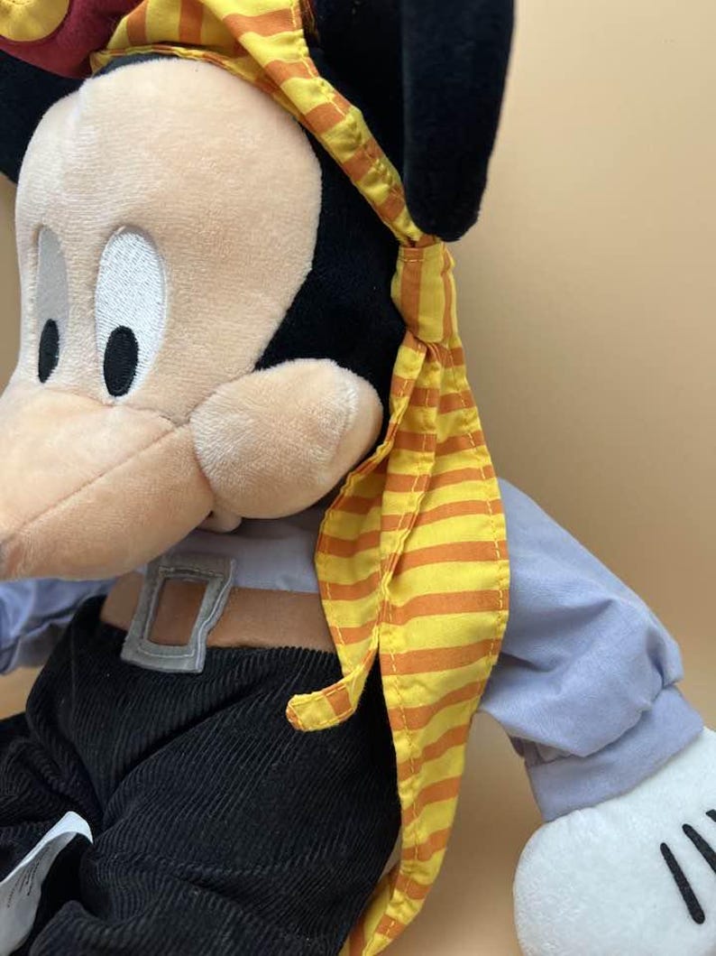 May include: A plush Mickey Mouse toy dressed as a pirate. The toy has a tan face, black ears, and is wearing a yellow and orange striped bandana. It is wearing a blue shirt, black pants, and a brown belt.