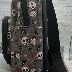 Puede incluir: Bolso bandolera negro y gris con un estampado repetido de caras, corazones y remolinos de Jack Skellington. El bolso tiene una correa negra ajustable, un bolsillo delantero y un cierre de cremallera. El bolso est&aacute; hecho de tela duradera.