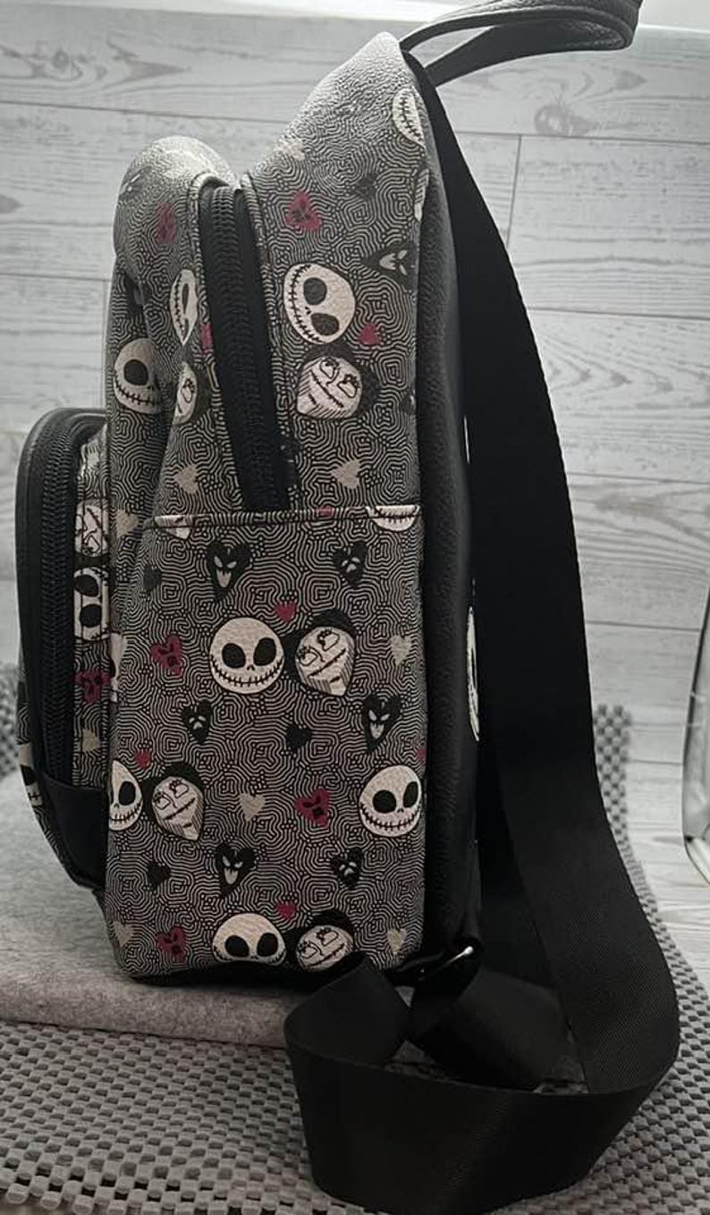 Puede incluir: Bolso bandolera negro y gris con un estampado repetido de caras, corazones y remolinos de Jack Skellington. El bolso tiene una correa negra ajustable, un bolsillo delantero y un cierre de cremallera. El bolso est&aacute; hecho de tela duradera.