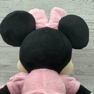 May include: A plush Minnie Mouse doll, viewed from the back. The doll features large black ears, a pink bow, and a pink dress. The arms and hands are black and white. The doll is made of soft, plush material.