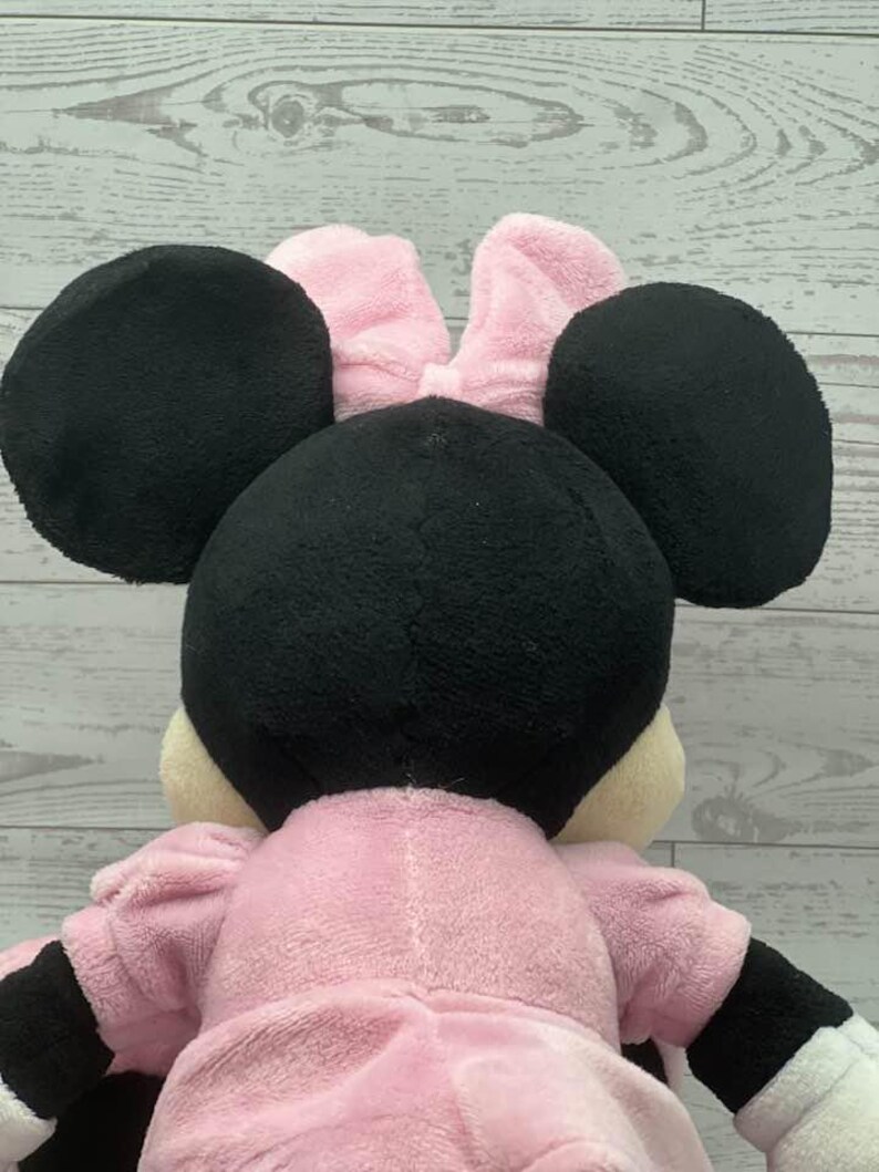 May include: A plush Minnie Mouse doll, viewed from the back. The doll features large black ears, a pink bow, and a pink dress. The arms and hands are black and white. The doll is made of soft, plush material.