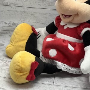 May include: A plush Minnie Mouse doll, wearing a red dress with white polka dots and a white lace trim. The doll has yellow shoes with red bows and black arms and legs. The doll's face is beige with black eyes and a black nose.
