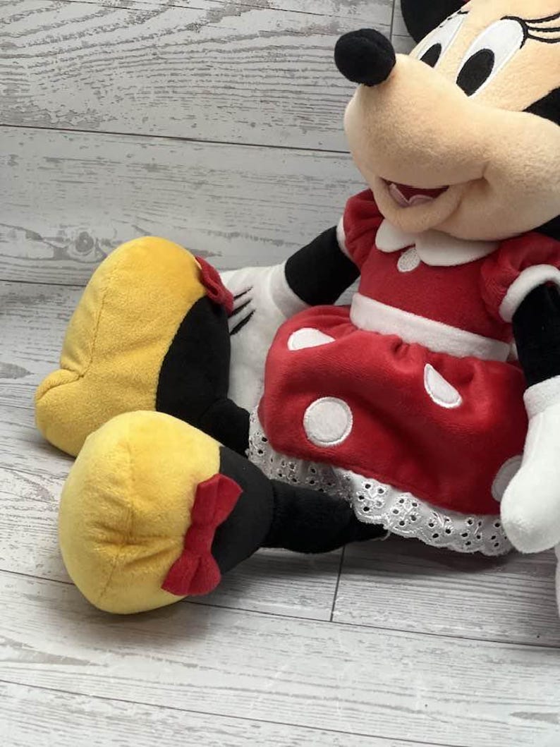 May include: A plush Minnie Mouse doll, wearing a red dress with white polka dots and a white lace trim. The doll has yellow shoes with red bows and black arms and legs. The doll's face is beige with black eyes and a black nose.