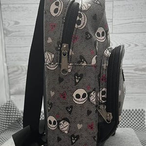 Puede incluir: Una mochila gris con un patr&oacute;n repetido de caras de Jack Skellington, corazones y remolinos. La mochila tiene cremalleras, correas y un peque&ntilde;o bolsillo delantero negros. La bolsa mide aproximadamente 30 cm de alto.