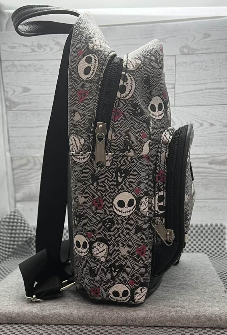 Puede incluir: Una mochila gris con un patr&oacute;n repetido de caras de Jack Skellington, corazones y remolinos. La mochila tiene cremalleras, correas y un peque&ntilde;o bolsillo delantero negros. La bolsa mide aproximadamente 30 cm de alto.