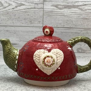 May include: A red ceramic teapot with a white heart design and a green handle and spout. The teapot has a textured appearance and a heart-shaped lid finial. The base is white, and the handle and spout have a knitted texture.