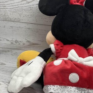 May include: A plush Minnie Mouse doll, with a black head, large ears, and a red bow. She is dressed in a red dress with white polka dots and white lace trim. A white gloved hand is visible.