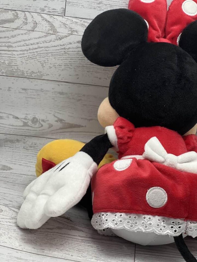 May include: A plush Minnie Mouse doll, with a black head, large ears, and a red bow. She is dressed in a red dress with white polka dots and white lace trim. A white gloved hand is visible.