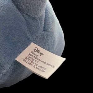 Puede incluir: Primer plano de un peluche azul claro con una etiqueta que dice "Disney" y "Fabricado bajo licencia por Just Play, LLC". El peluche est&aacute; hecho de un material suave y difuso.