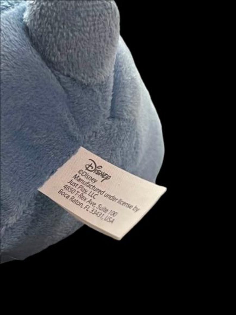 Puede incluir: Primer plano de un peluche azul claro con una etiqueta que dice "Disney" y "Fabricado bajo licencia por Just Play, LLC". El peluche est&aacute; hecho de un material suave y difuso.