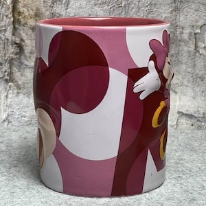 Op de afbeelding: Een cilindervormige keramische mok met een roze binnenkant en een buitenkant met een abstract cirkelpatroon in verschillende tinten roze en bordeauxrood. De mok heeft een reli&euml;f van een stripfiguur met een roze strik en witte handschoenen.